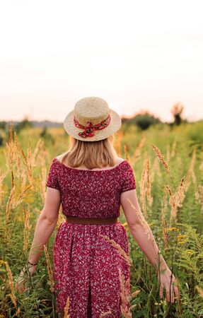 Beautiful girl in red dress in yellow walking through field, sunset time. golden hour. village sceneryの写真素材