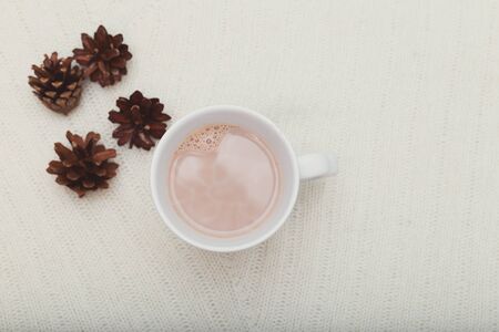 A cup of cocoa stands on a white knitted blanket, beside lie cones