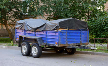 A heavy four-wheeled cargo trailer with a dark awning is parked near the green trees
