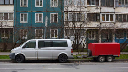 A minivan with a four-wheeled cargo trailer is parked at a residential building
