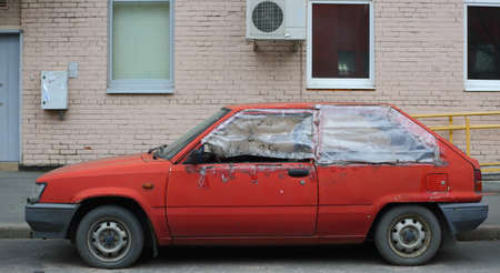 An old wrecked red hatchback car is parked outside the house