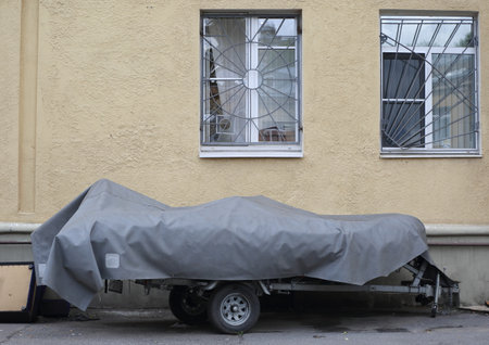 Covered with a gray cover, a long two-wheeled trailer is parked at the yellow wall, ulitsa Sedova, St. Petersburg, Russia, August 2021のeditorial素材