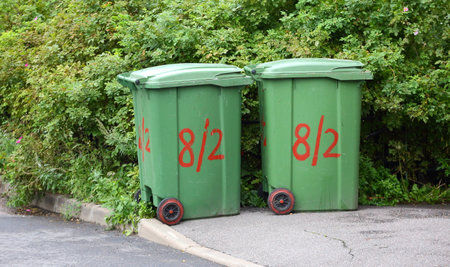 Two small green plastic mobile dumpsters are parked near the bushesの写真素材