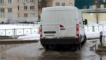 A white minibus rides through a puddle during the thaw season