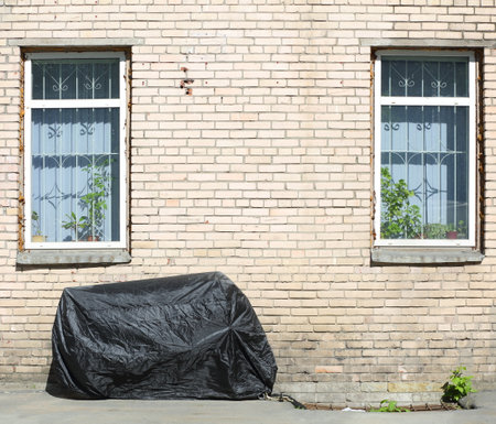 A motorcycle, covered with a cover from bad weather, stands near a white brick wall between two windowsの写真素材