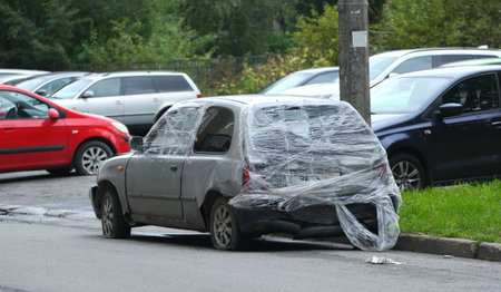 An old gray broken-down car parked on the street is covered with a transparent plastic filmの写真素材