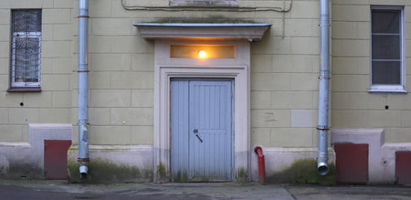 The wall of an old house with narrow windows, drainpipes, and a burning lamp above the wooden front...