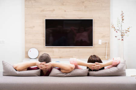 Back view of couple man and woman resting in apartment, sitting on sofa together and watching TVの写真素材