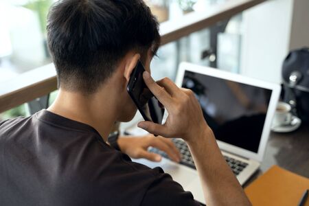 Young handsome businessman working with laptop at desk in the coffeeshop, talking on phone, have good planning and organizational skills, making good annual income, promoting and marketing businessの写真素材