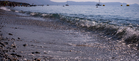 a foamy wave rolls onto a pebble sea beach with boats in the distance
