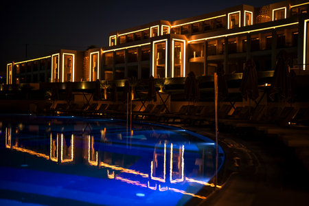 Illuminated modern architectural structures of luxury hotel reflected in huge blue swimming pool at nightの写真素材