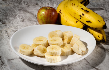 A bunch of bananas, a sliced banana, and an apple on a plate on a table. Selective focus.の写真素材