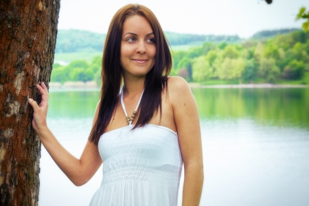 Beautiful woman standing near on a tree, coast of a lake in the backgroundの写真素材