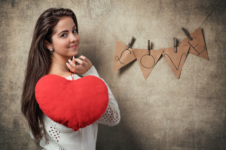 Valentines Day / Young woman standing before a rustic background with heart shaped pillow and text LOVEの写真素材