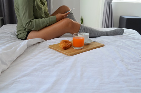 Breakfast in bed, beautiful woman is sitting on the bed, she holds a phone in her hand.の写真素材
