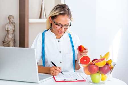 Smiling nutritionist in her office, she is holding a green apple and showing healthy vegetables and fruits, healthcare and diet concept. Female nutritionist with fruits working at her deskの写真素材