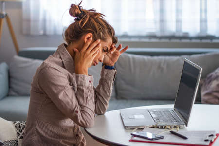 Young frustrated woman working at office desk in front of laptop suffering from chronic daily...