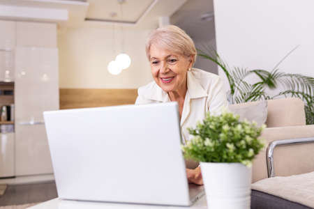 Portrait of senior woman sitting at table at home and working on her laptop. Older lady surfing the...