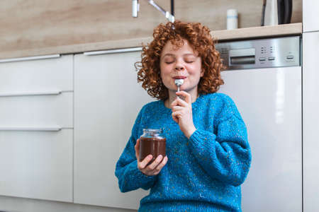 young woman eating chocolate from a jar while sitting on the wooden kitchen floor. Cute ginger girl indulging cheeky face eating chocolate spread from jar using spoon savoring every mouthfulの写真素材
