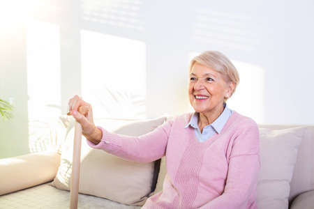 Retired woman with her wooden walking stick at home. Happy senior woman relaxing at home holding cane and looking at camera. Smiling grandmother sitting on couch.の写真素材