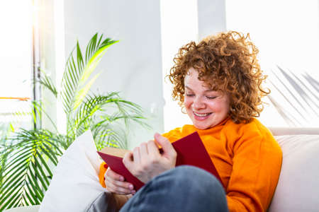 Young woman at home sitting on modern sofa relaxing in her living room reading book . White cozy bed...
