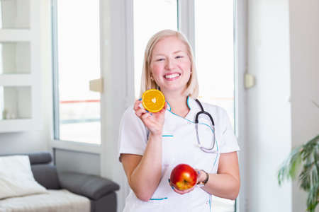 Smiling nutritionist in her office, she is holding a fruit and showing healthy vegetables and fruits, healthcare and diet concept. Female nutritionist with fruitsの写真素材