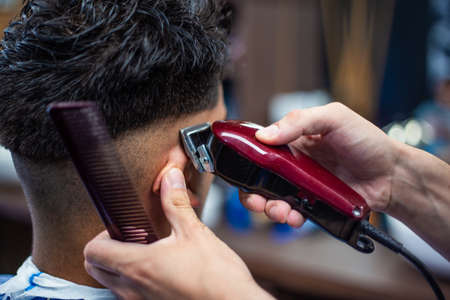 Close up shot of man getting trendy haircut at barber shop. Male hairstylist serving client, making haircut using machine and comb.の写真素材