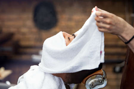 Barber covers the face of a man with a hot towel. Traditional ritual of shaving the beard with hot and cold compresses in a old style barber shop. Client with hot towel on face before shaving in barber shopの写真素材