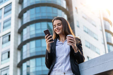 Business woman with coffe and talking on the phone near office. Young woman with smartphone standing...