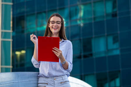 Happy beautiful business woman with clipboard. Portrait Of Successful Business Woman Holding clipboard. Photo business woman wearing suit and holding documents in hands. Panoramic windows background.の写真素材