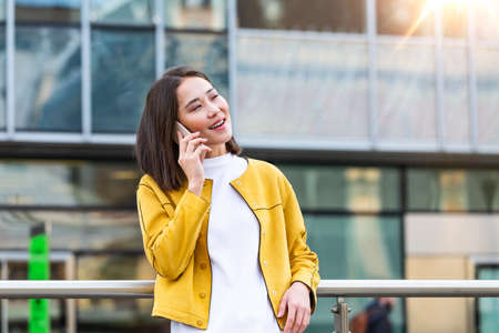 Happy smiling business woman talking on mobile smartphone in the street with office buildings in the background. Executive business Asian woman talking on mobile smartphoneの写真素材