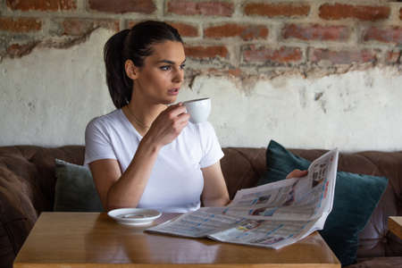 Charming woman with beautiful smile reading newspaper during rest in coffee shop, happy Caucasian...