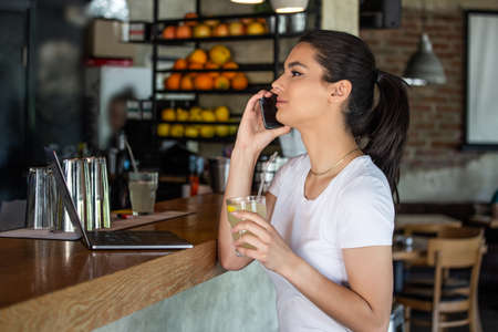 Young charming woman calling with cell telephone while sitting alone in coffee shop during free time, attractive female with cute smile having talking conversation with mobile phone while rest in barの写真素材