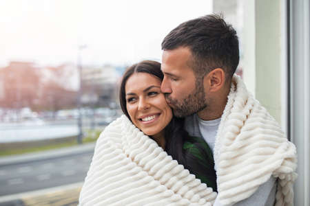 Romantic couple on a winter holiday. Man and woman standing together in a hotel room balcony wrapped in blanket. Couple embracing and smiling.の写真素材