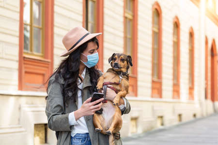 Woman during pandemic isolation walking with her dog and using mobile phone on the street. Stylish girl with funny dog resting, hugging and having fun, cute moments.の写真素材