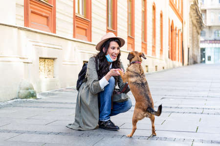 Young woman with long hair and face protective mask embracing funny dog on the street during coronavirus pandemic isolation. Smiling young woman enjoying good day and posing with pet.の写真素材