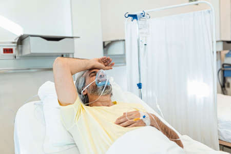 Male coronavirus patient looking away while resting on hospital bed. Man is wearing oxygen mask. He...