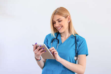 Young female clinician doctor in scrubs using touchpad while communicating with patients online. Portrait Of Smiling Female Doctor Wearing Scrubs In Hospital Corridor Holding Digital Tabletの写真素材