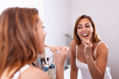 Portrait of attractive woman brushing teeth in bathroom and looking in the mirror at reflection....