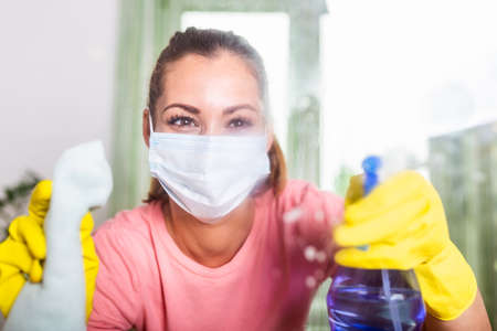 Woman with protective glove and facial mask doing housework, housewife portrait. She sprinkling...