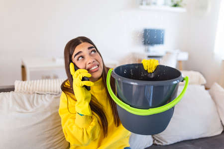 Worried Young woman Calling Plumber While Leakage Water Falling Into Bucket At Home
