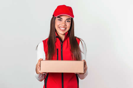 Image of a happy young delivery woman in red cap standing with parcel post box isolated over white background.の写真素材