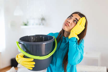 Shocked Woman Looks at the Ceiling While Collecting Water Which Leaks in the Living Room at Home. Worried Woman Holding Bucket While Water Droplets Leak From Ceiling in Living Roomの写真素材