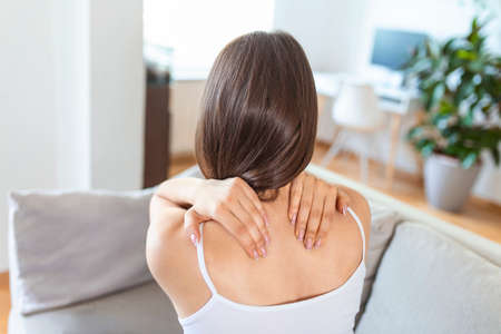 Young woman suffering from backache at home. Portrait of a young brunette girl sitting on the couch at home with a neckache and back pain. Beautiful woman Having Spinal Or neck painの写真素材