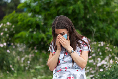 Portrait of beautiful sad woman in medical mask sneezes among white flowers. coronavirus pandemic. Concept of not spread of the virus.の写真素材