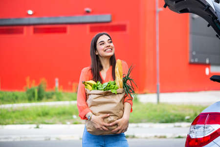 Young woman riding shopping cart full of food on the outdoor parking. Young woman in car park, loading shopping into boot of car. Shopping successfully done. Woman putting bags into car after shoppingの写真素材