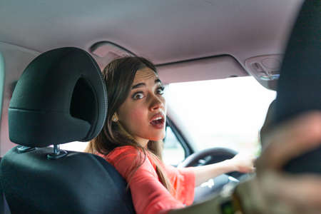 Photo of a woman in car indoor keeps wheel, turning around looking at passengers in back seat idea taxi driver. Concept of exam Vehicle. Back view of an attractive young businesswoman looking over her shoulder while driving a car.の写真素材