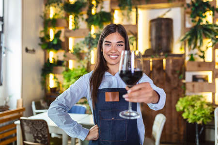 Young beautiful waitress wearing apron holding a glass of red wine in one hand serving a customer in a rustic restaurant. Sommelier recommended wine in restaurant.の写真素材