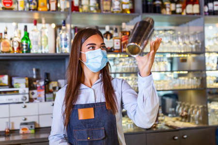 Young female worker at bartender desk in restaurant bar preparing coctail with shaker. Beautiful young woman behind bar wearing protective face maskの写真素材