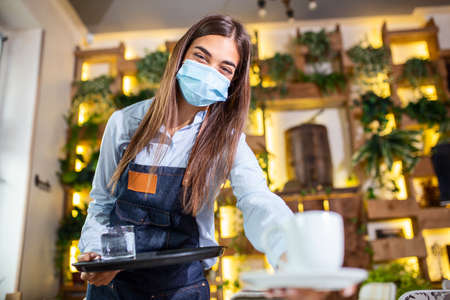 Female waitress in a medical protective mask serves the coffee in restaurant durin coronavirus pandemic representing new normal conceptの写真素材